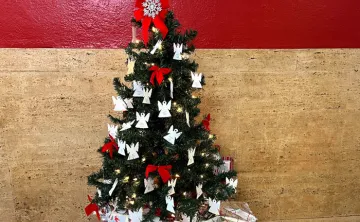 A small Christmas tree with paper angel ornaments and red bows in front of red plus tan stone wall.
