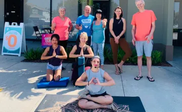 YMCA staff and members smiling outside new facility. Three people are on the ground in yoga pose with prayer hands and five others stand behind them.