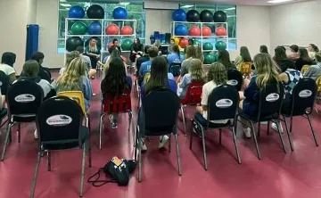 Teens in a large room with red floors, sitting in chairs listening to adult speaker during a meeting.