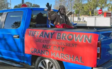 Ridgecrest leader waves from the bed of a blue truck for Greater Ridgecrest MLK Day parade. Sign is on side of truck bed, black block letters in all caps: Tiffany Brown Greater Ridgecrest MLK Grand Marshal on red background.