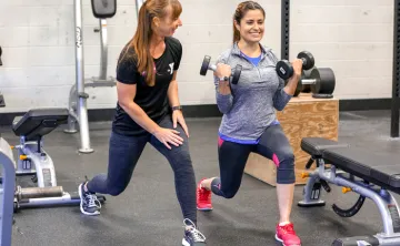 Female personal trainer demonstrates a forward lunge to female client, as client is in the stance. Client is holding dumbbells in bicep curl position.