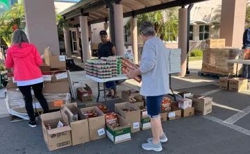Four volunteers at James P. Gills YMCA organize food donations outside in the shade. Many brown boxes of food rest on pallets and the ground. Foreground includes a man facing away from the camera, holding cereal boxes.
