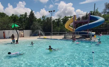 Kids playing in outdoor zero-entry pool with waterslides in the background. Sunny day with puffy white clouds and tall evergreen trees in background scenery.