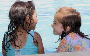 Two toddlers with wet hair and smile at each other on pool ledge during water safety class.