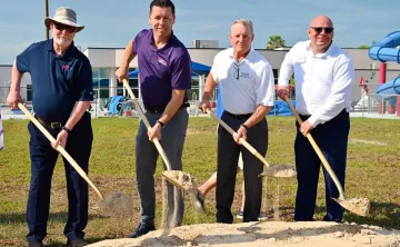 Outdoor image of four men posing for a groundbreaking photo. The subjects are holding shovels with dirt. A swimming pool with slides is in the background.