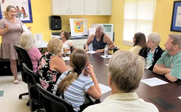 A group of nine adults sitting at a table, listening to a woman standing, teaching the class. Background: classroom walls are yellow.