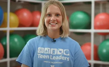 A teen girl with short blonde hair and a light blue Teen Leaders YMCA shirt smile at the camera. The background is out of focus, and shows white shelves holding green and red exercise balls.