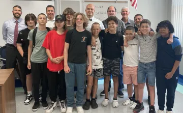 A group of five adults stand behind 10 middle school students, posing for a photo. The setting is a school classroom, a portion of the hanging American flag is shown in the background.