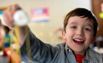 A young boy smiles as he reaches his arm out to show off the craft he made. The child's face is in focus and the craft is in the foreground, out of focus.