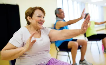 Three seniors holding arms out during chair exercises in fall prevention program. Woman wearing purple pants is in focus, two seniors in the background are out of focus.