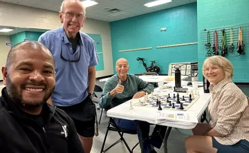 4 members of Chess Club smiling for photo. Two are sitting beside the chess table.
